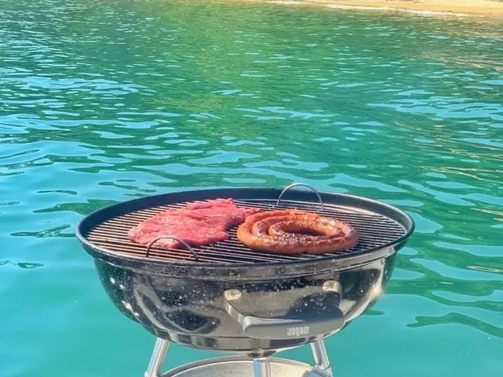 Churrasqueira portátil com linguiças e carnes, sobre o mar azul-turquesa de uma enseada
